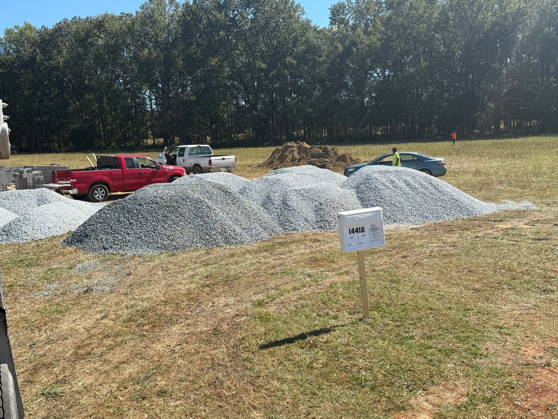 Piles of gravel on a grassy field, with trucks and workers in the background. Sunny day.