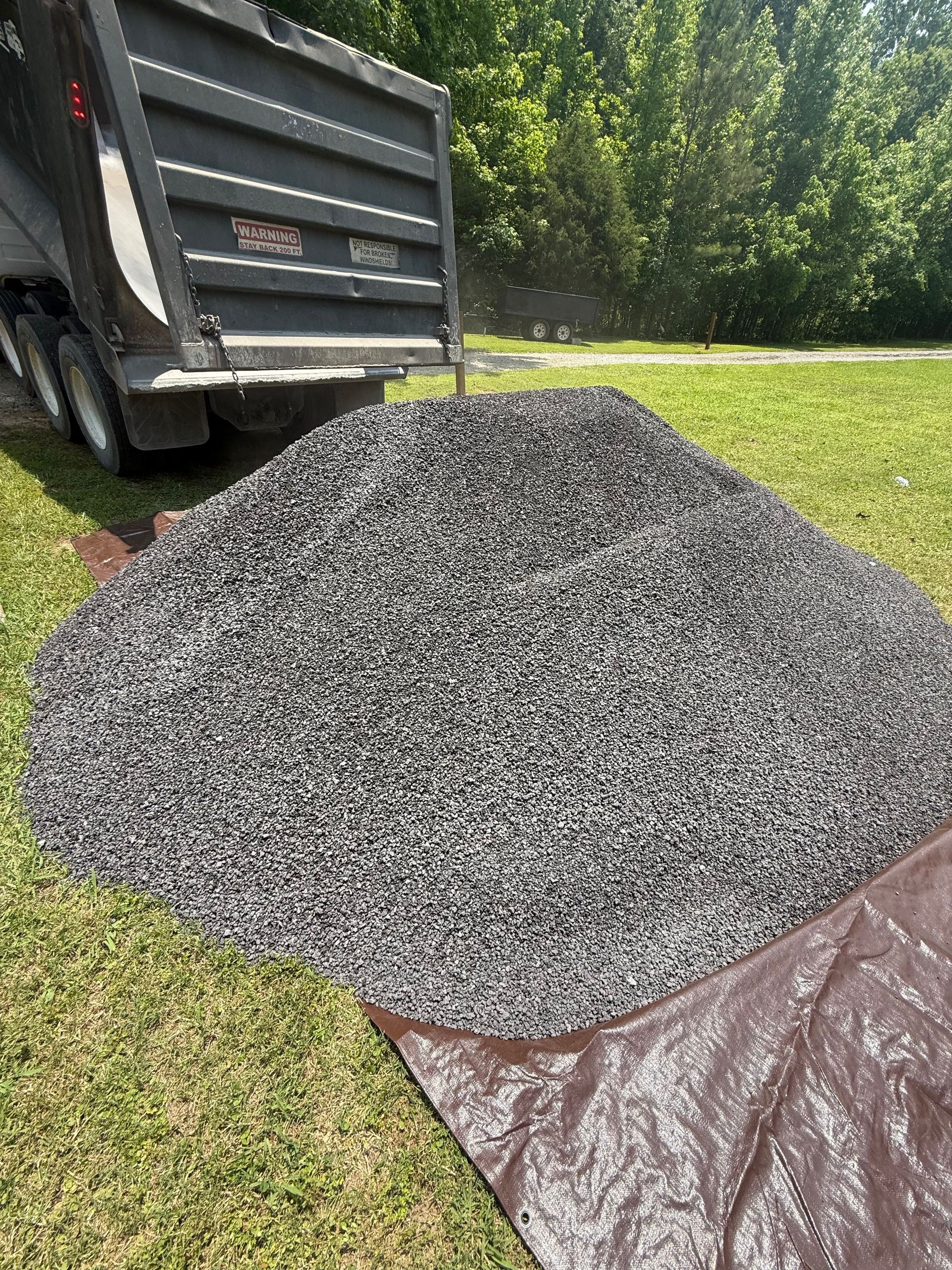 A dump truck unloading a large pile of dark gray gravel onto a brown tarp in a grassy yard.