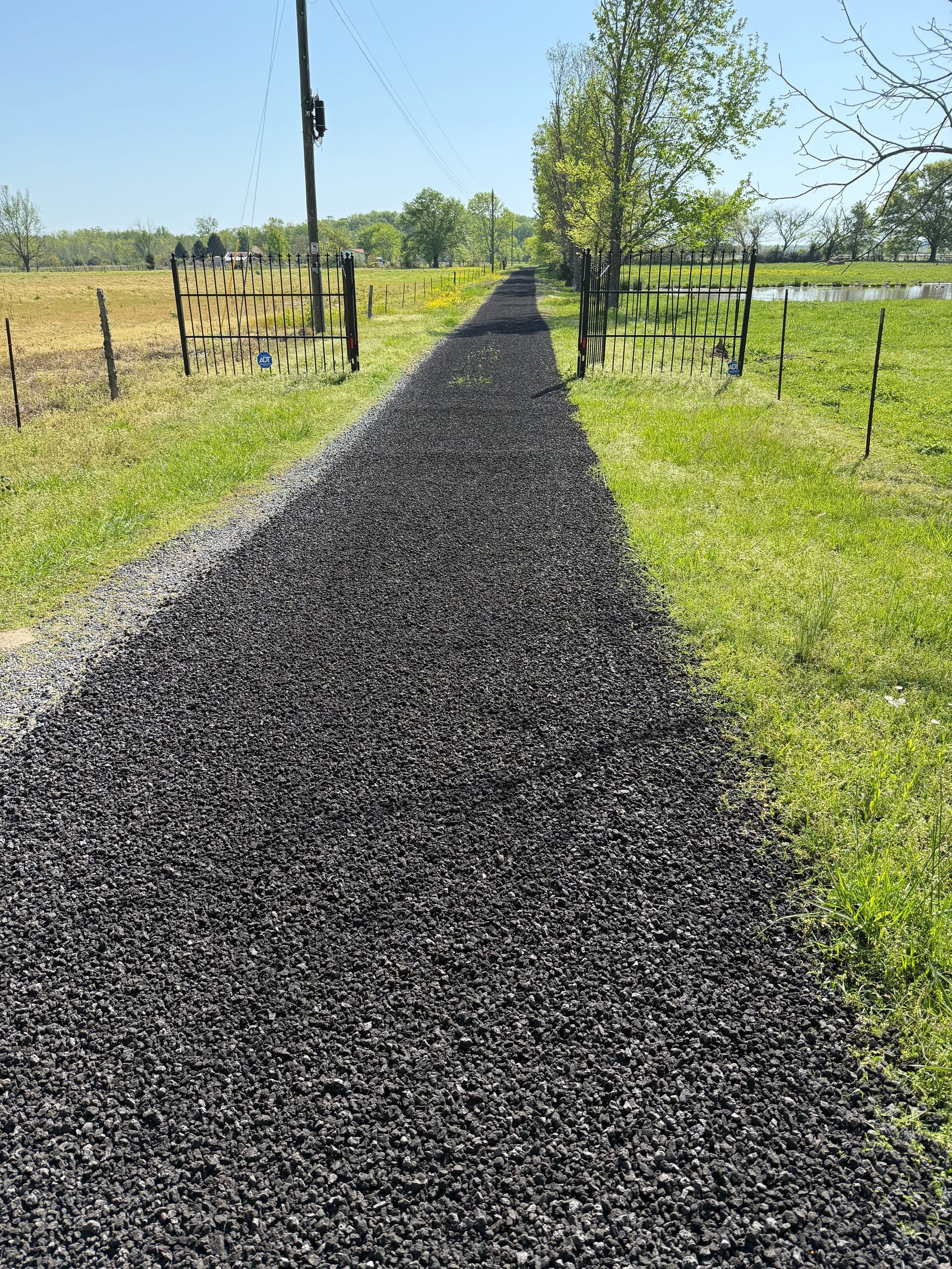 Black pathway through green grass and open gates, leading to a field.