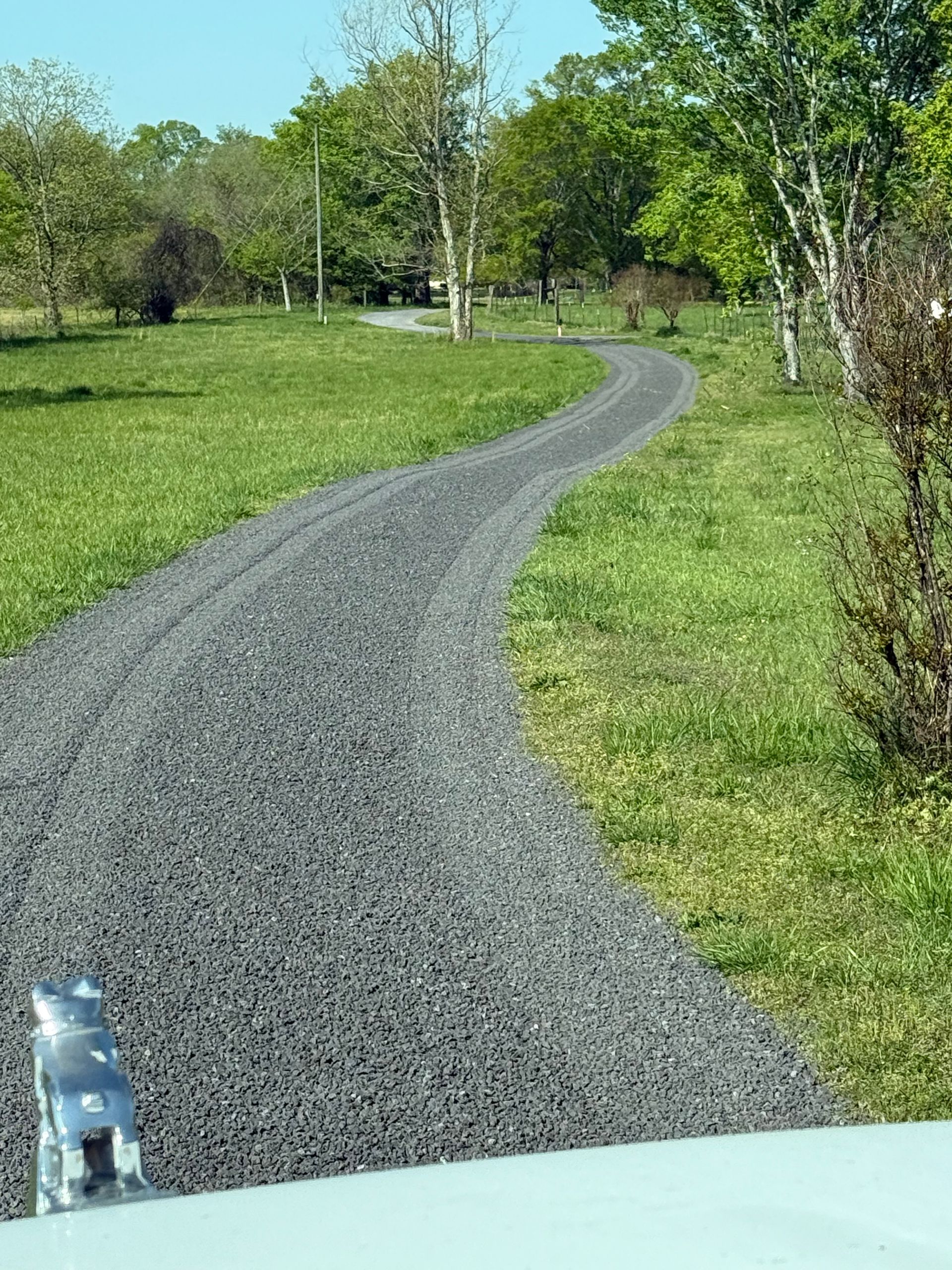 Gravel road winding through a grassy field, trees in the background, blue sky visible.