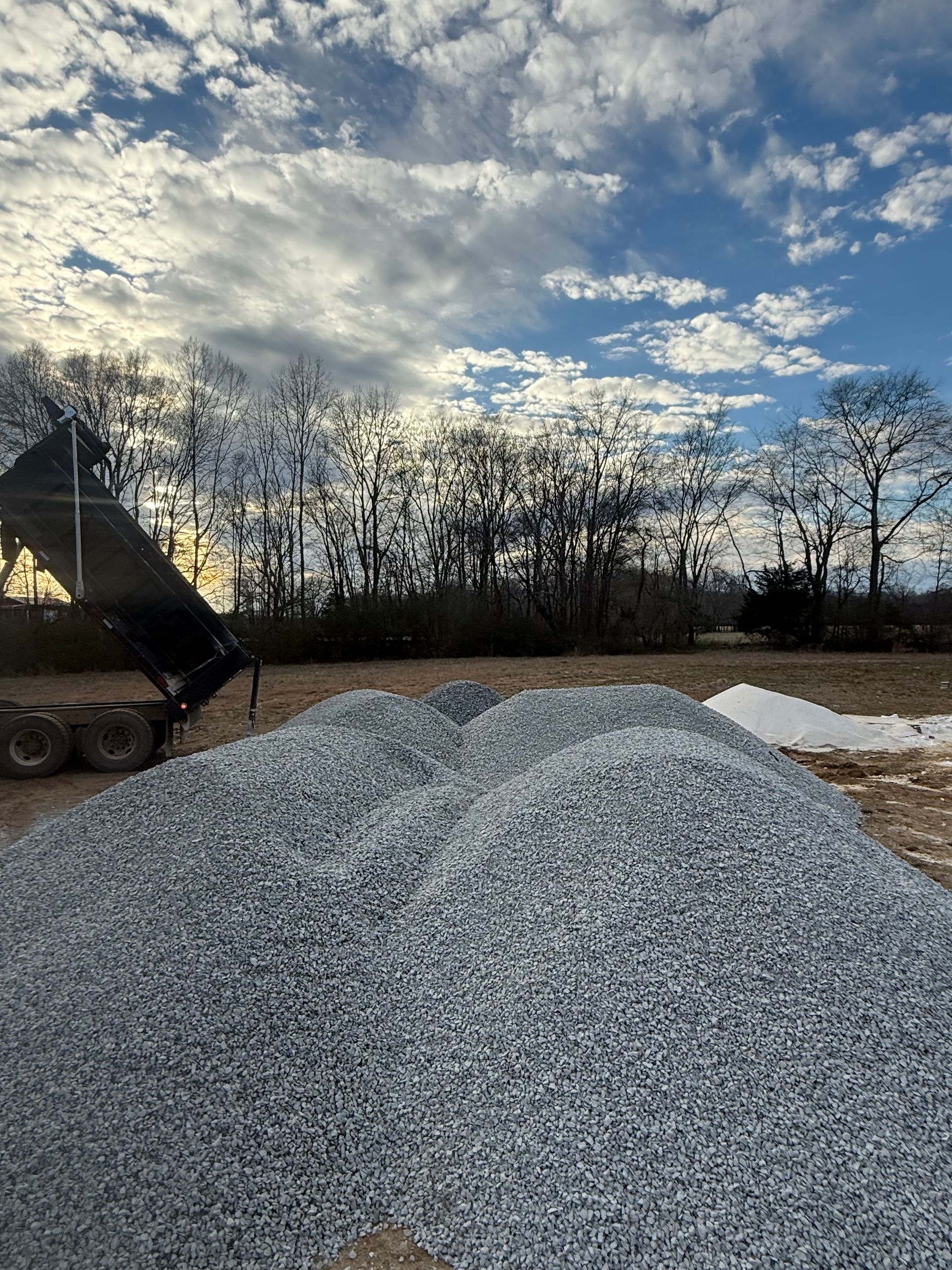 Dump truck dumping gravel onto a field under a cloudy sky.