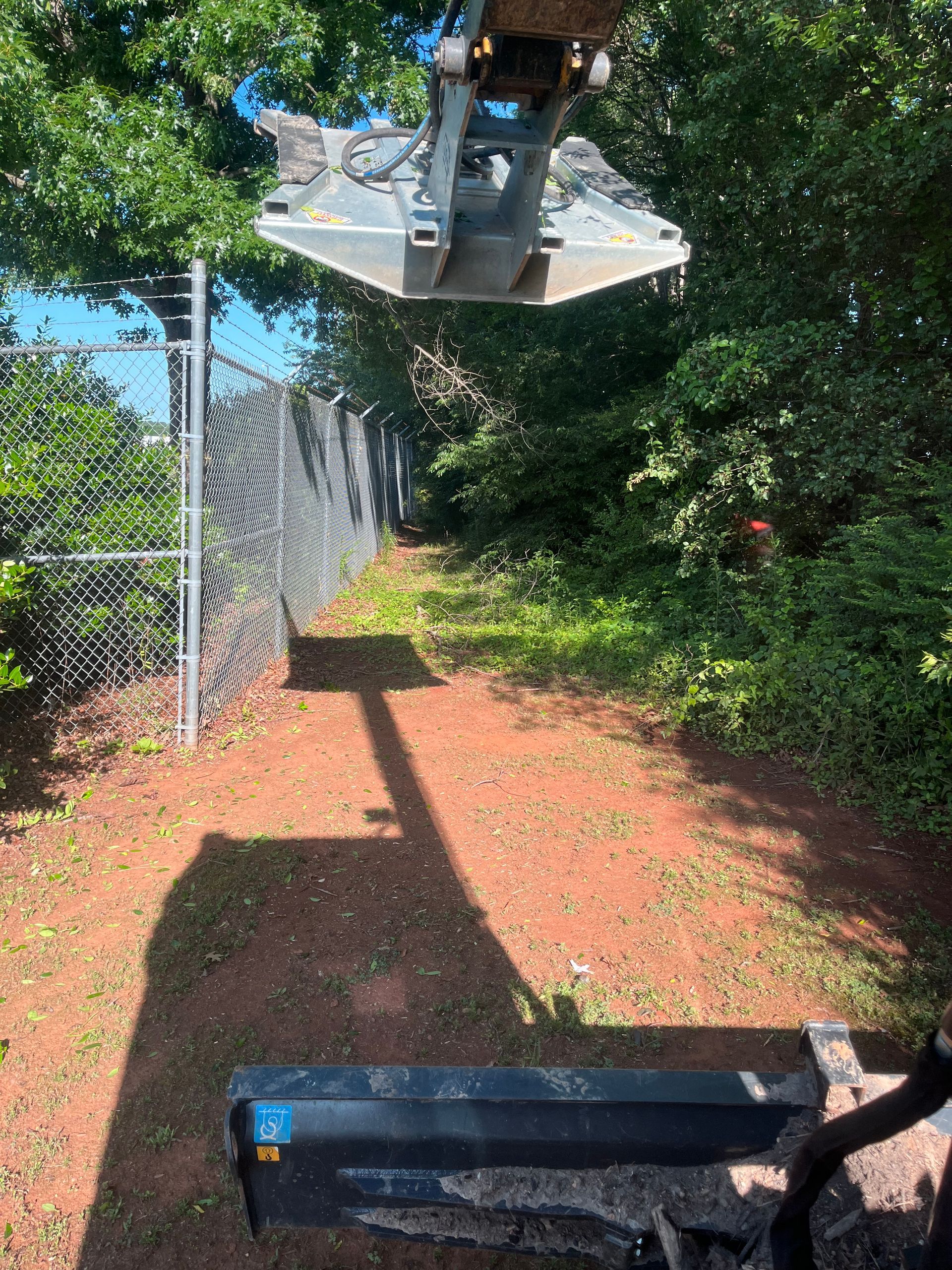 A shadow of a crane is cast on a dirt path next to a chain link fence.