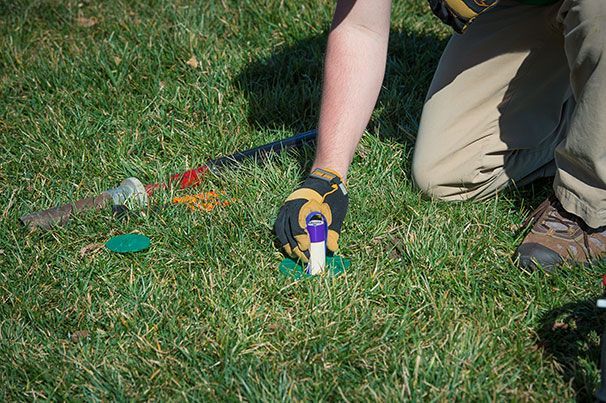 Person kneels on grass, installing a sprinkler head. Gloves, tan pants, and green grass are visible.