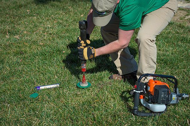 Person using a gas-powered auger in grass to drill a hole; a flag and tube are nearby.