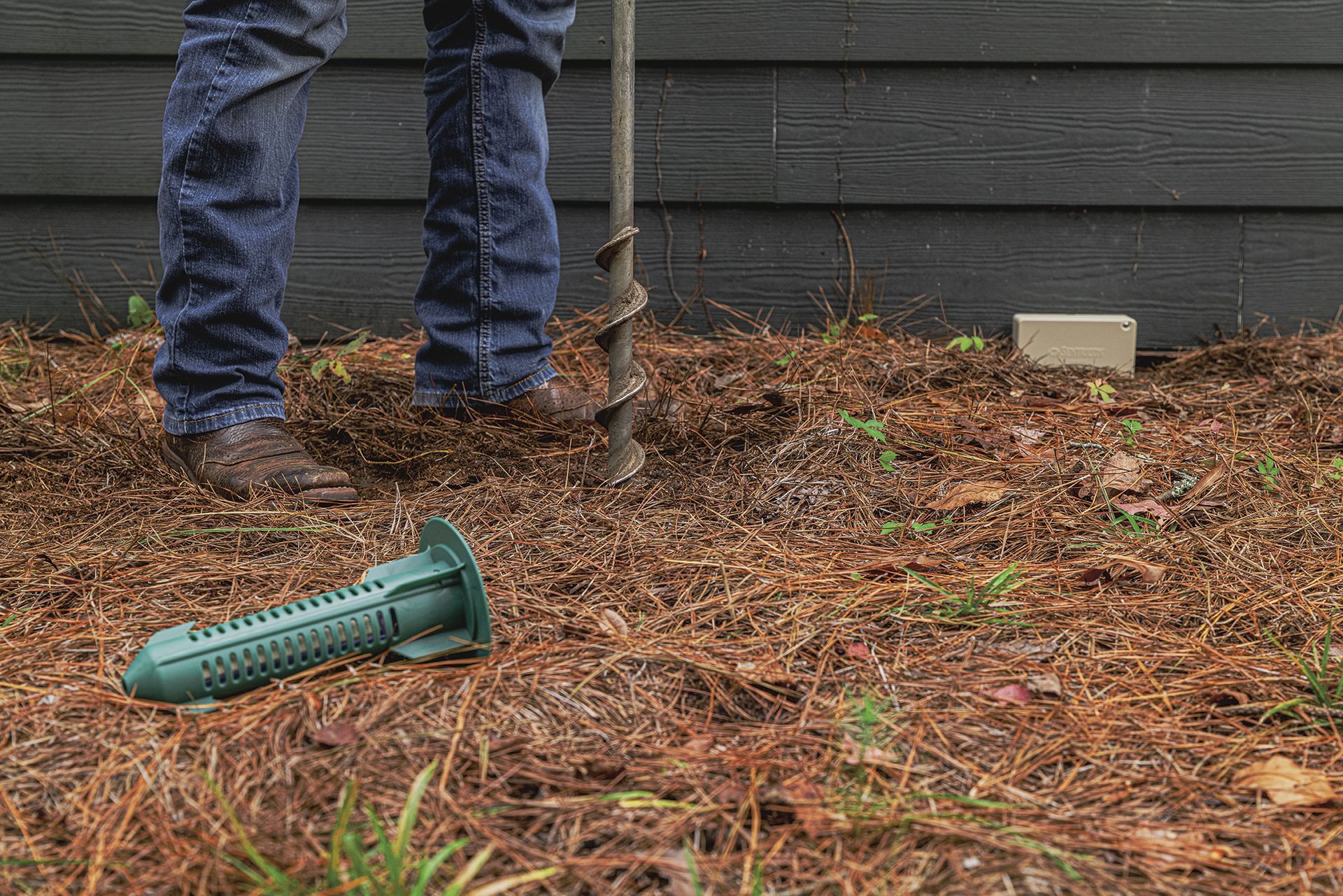 A person using a drill to install a green termite bait station near a house.