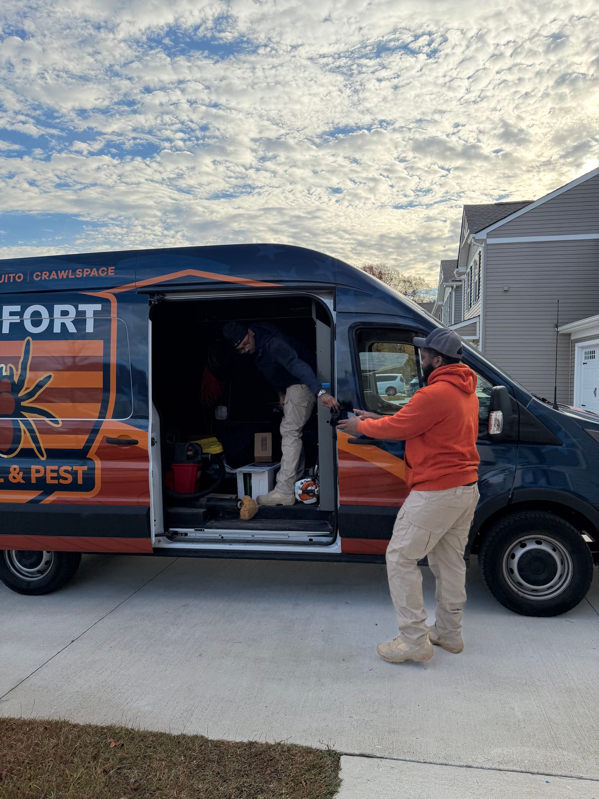 Two men exiting a pest control van in front of a house on a cloudy day. The van is navy and orange.