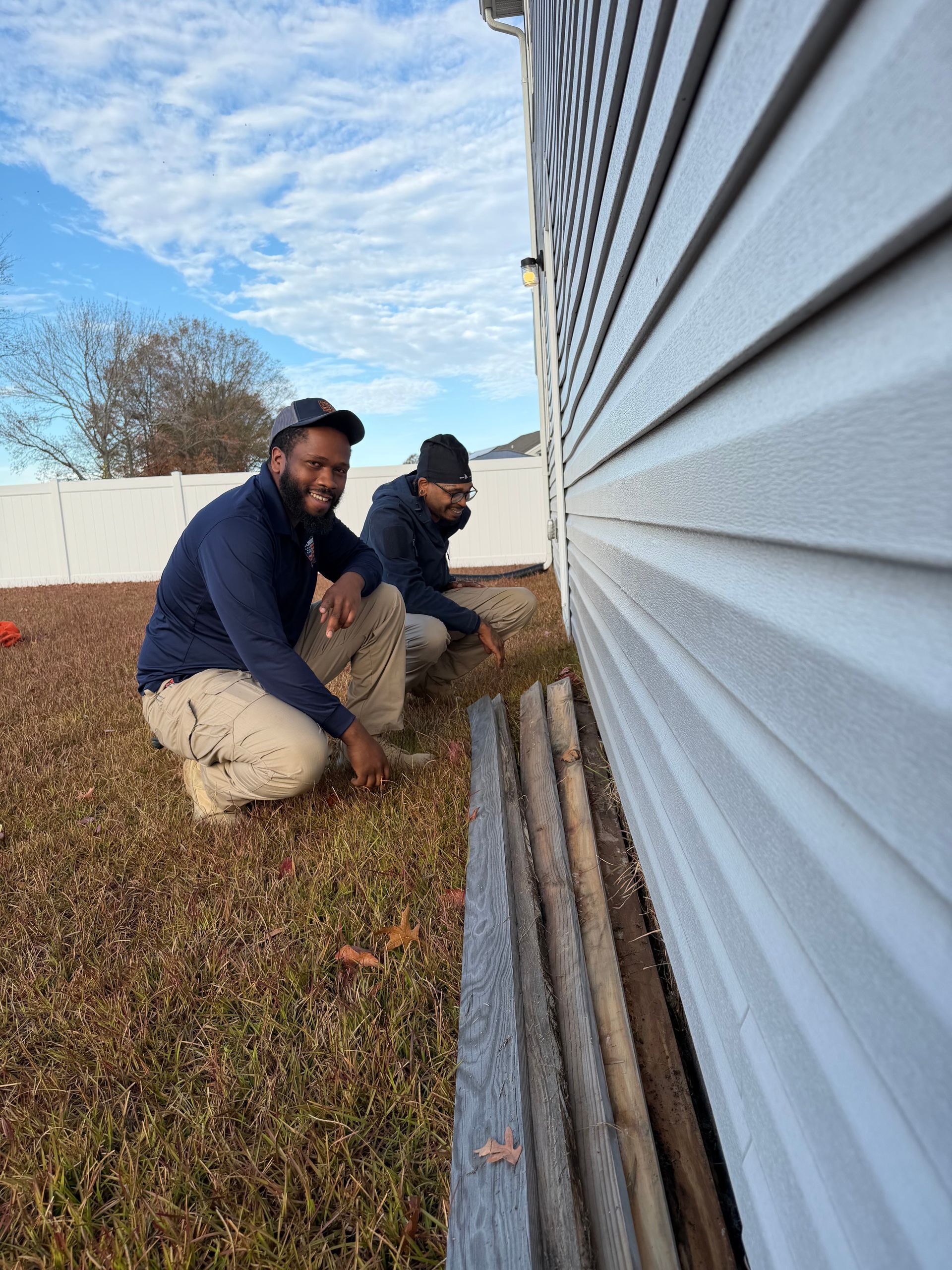 Two people squatting next to a building, examining the ground. One smiles. Blue sky, grass, and white siding.