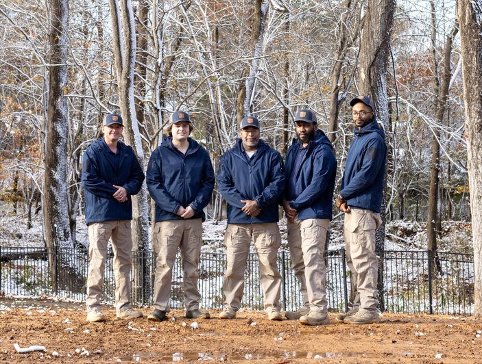 Five people in blue jackets and khaki pants stand outdoors in a snowy wooded area.
