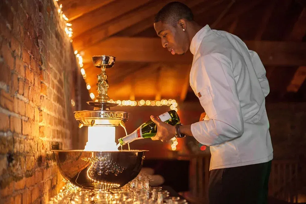 Man pouring champagne into a tiered fountain with glasses set out; warm lighting.