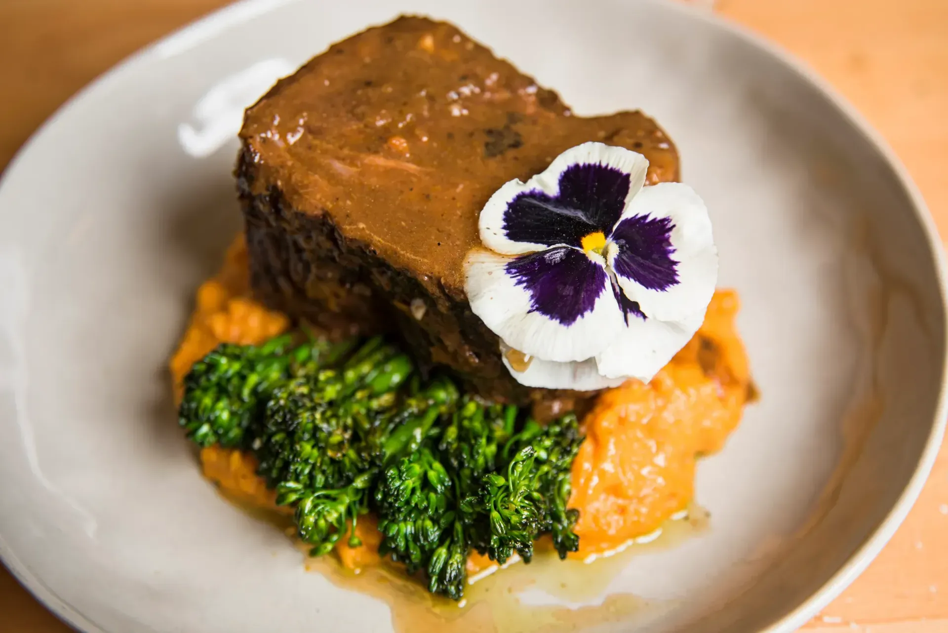 Braised meat with gravy, mashed sweet potato, and broccoli, garnished with a pansy.
