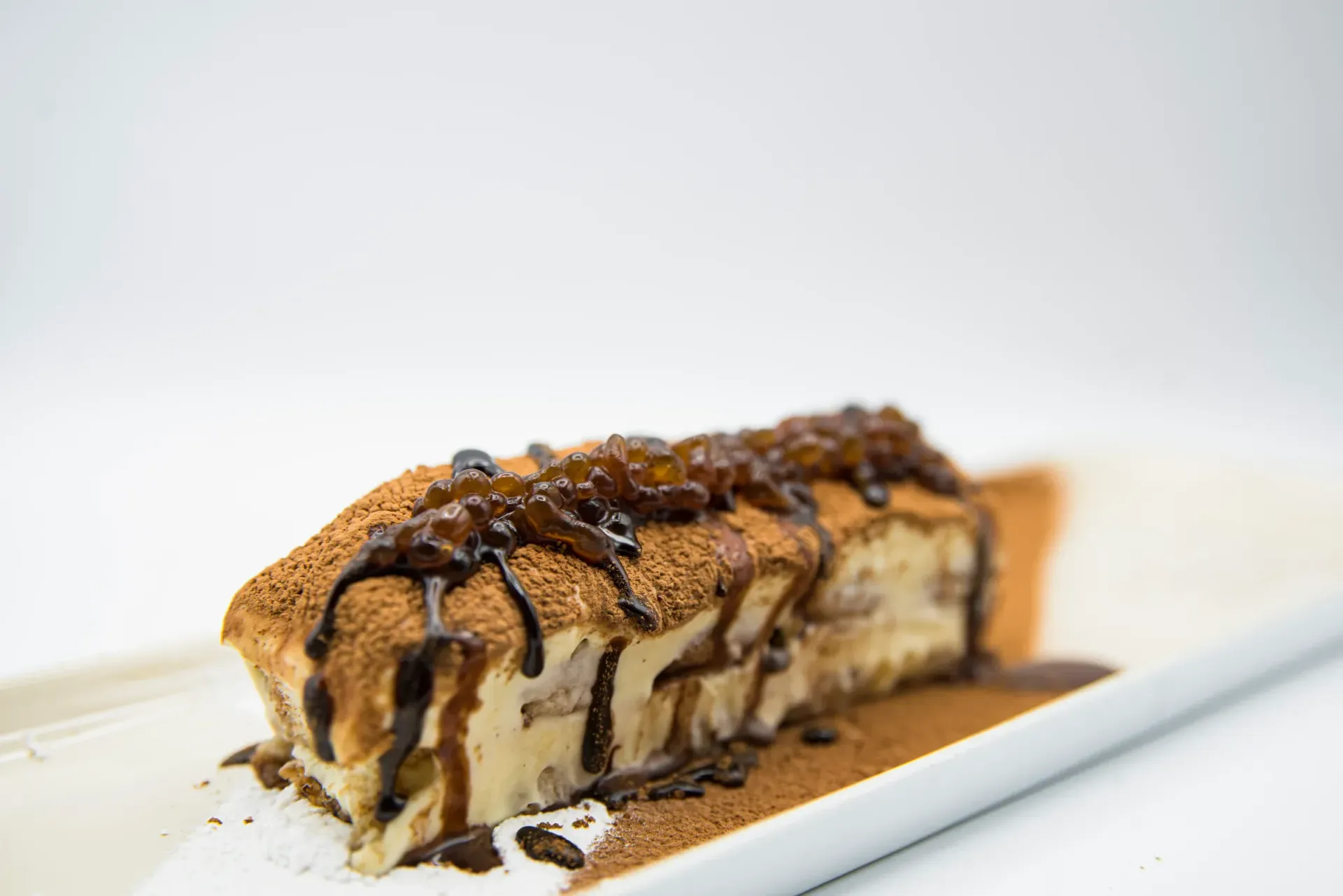 Tiramisu dessert on white plate, topped with chocolate syrup, cocoa powder, and dark caramel.