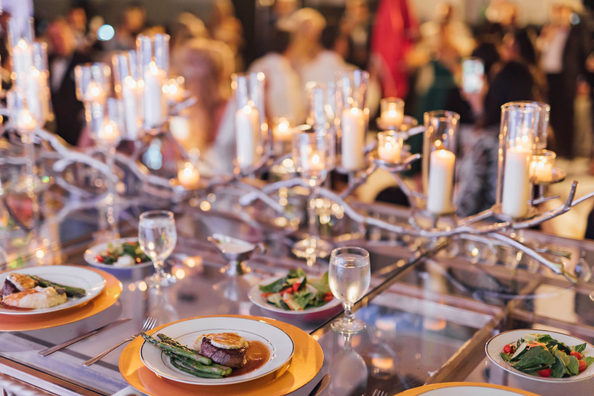 Elegant table setting with lit candles, food plates, glasses, and blurred background of people.