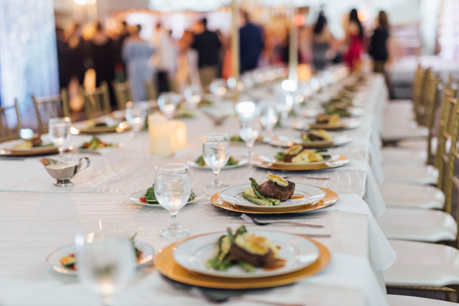 Long dining table set for a formal event, with plated meals, glasses, and blurred background of guests.