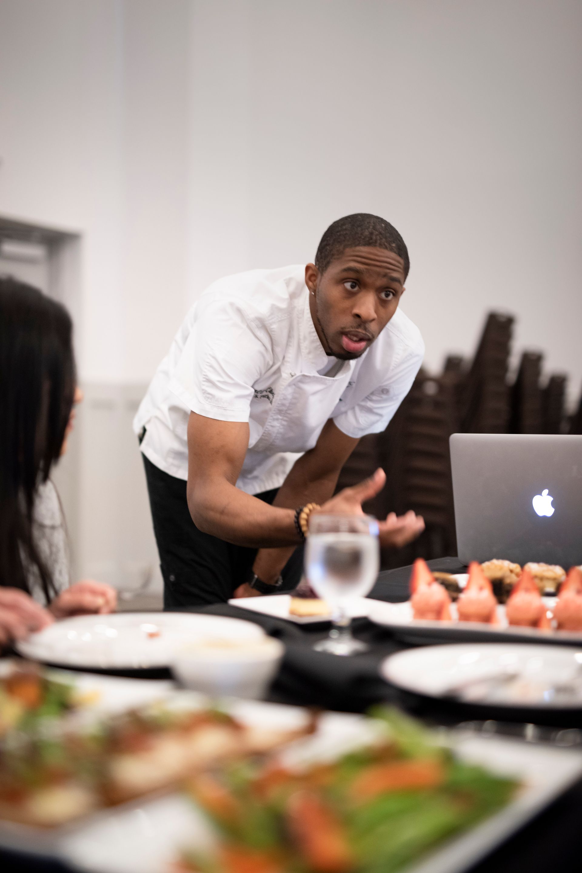 Chef gesturing while explaining something at a table setting, laptop and food present.