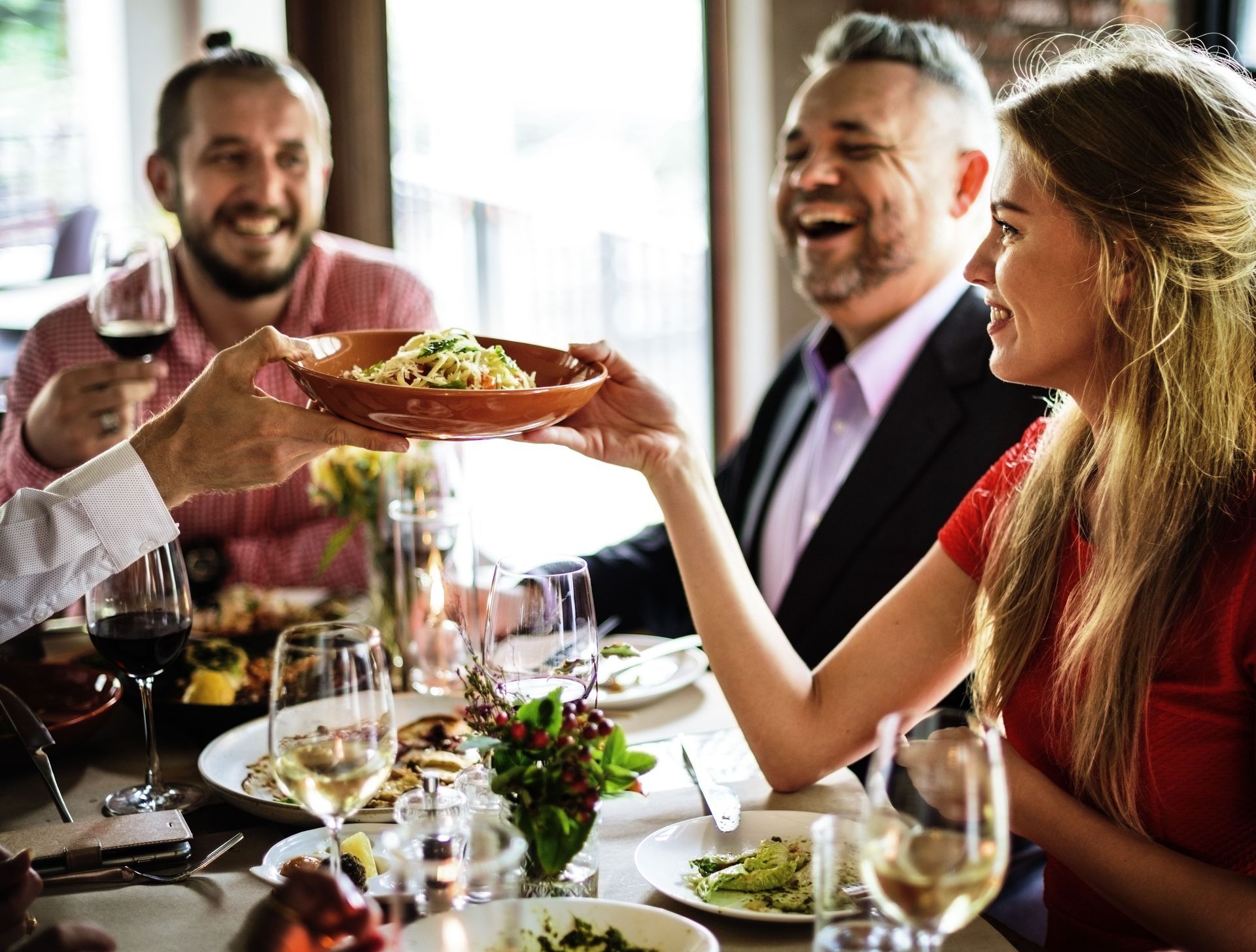 People sharing a meal at a restaurant, smiling and passing a dish across the table.