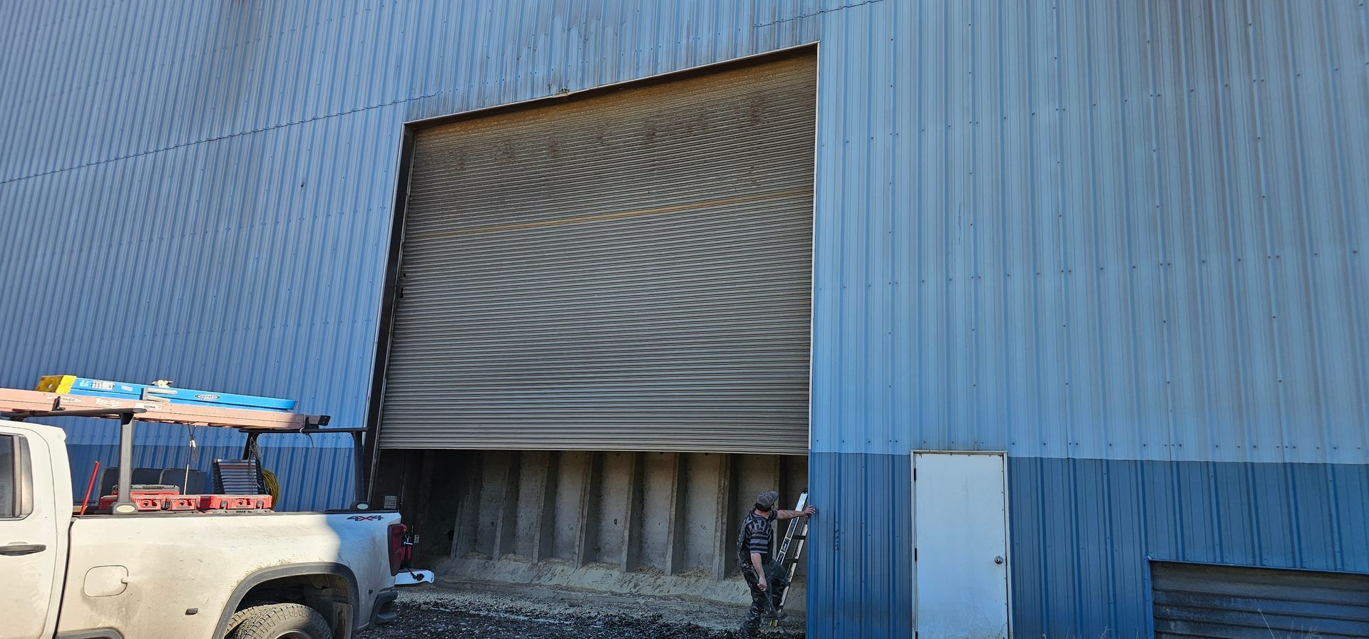 A white truck is parked in front of a blue building with a broken garage door.