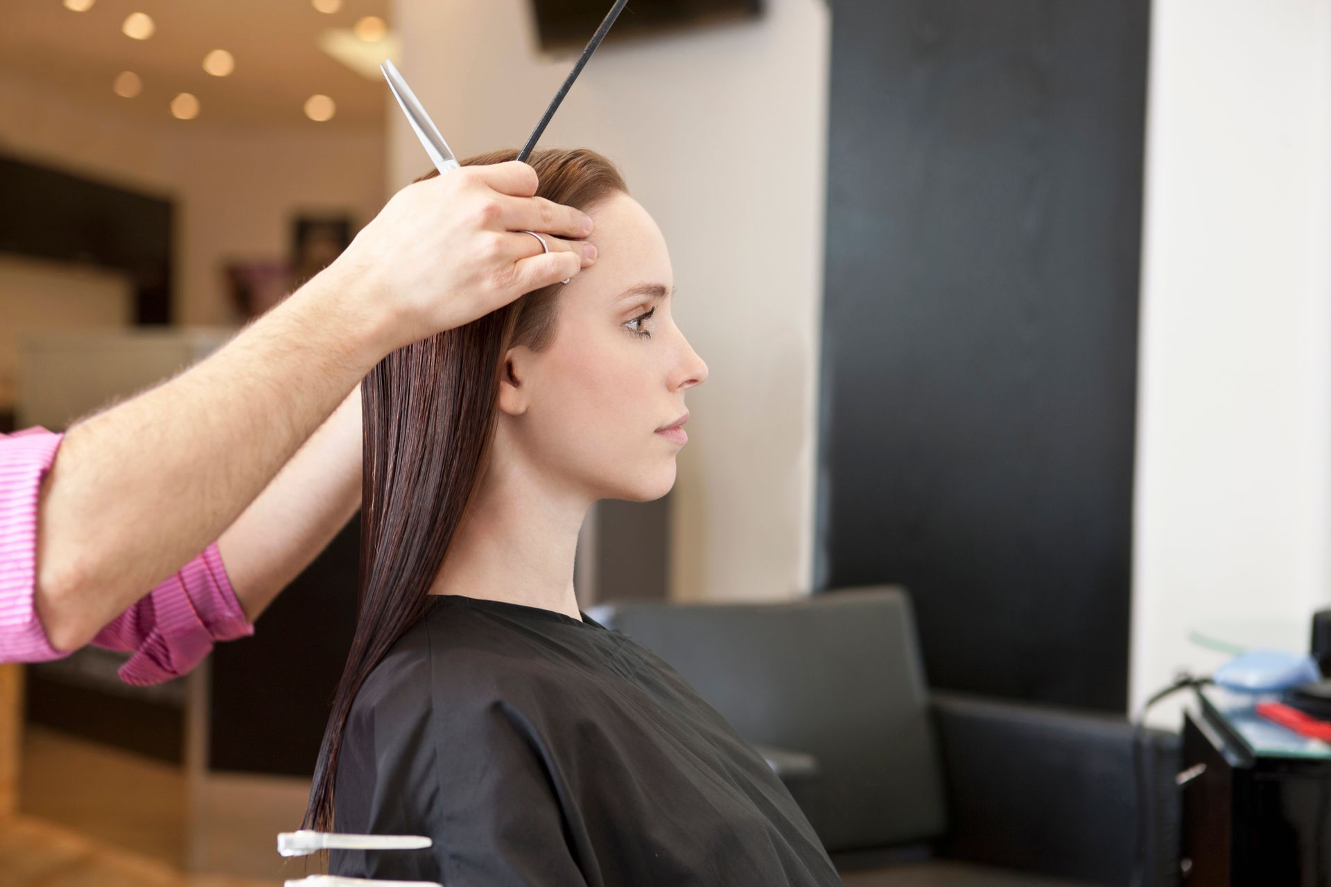 Hairdresser cutting a woman's long, wet hair at a salon. The woman wears a black cape, and the walls are neutral.