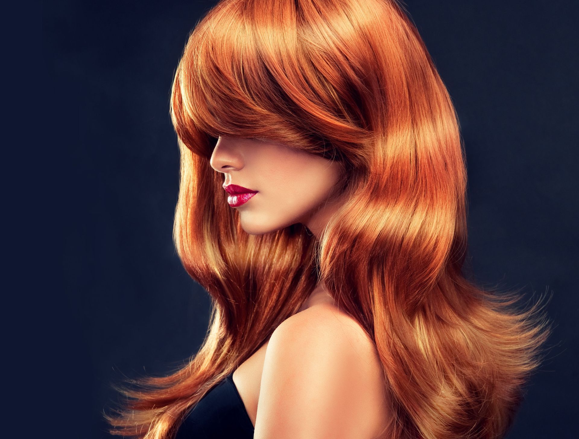 Woman with vibrant auburn hair, side profile, with a black top, red lipstick, against a dark background.