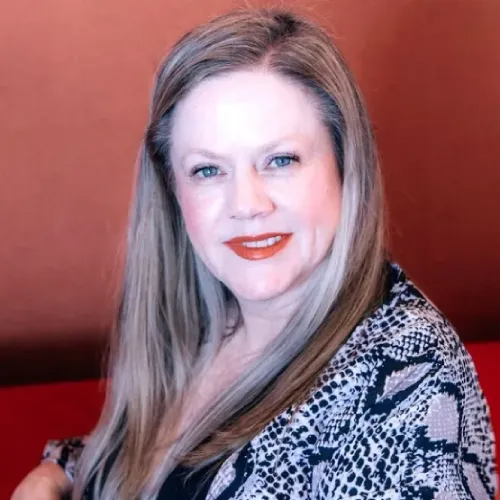 Michaelle Holland with long, light brown hair smiles; she wears a snake print jacket, sitting in front of a red wall.