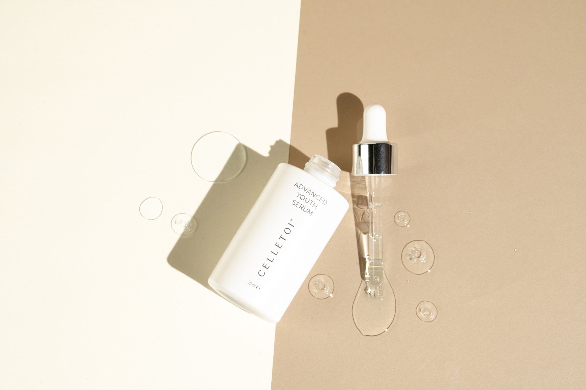 White skincare bottle with a dropper, and water droplets on a cream and beige background.