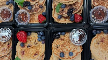 A bunch of pancakes with berries and syrup in plastic containers.