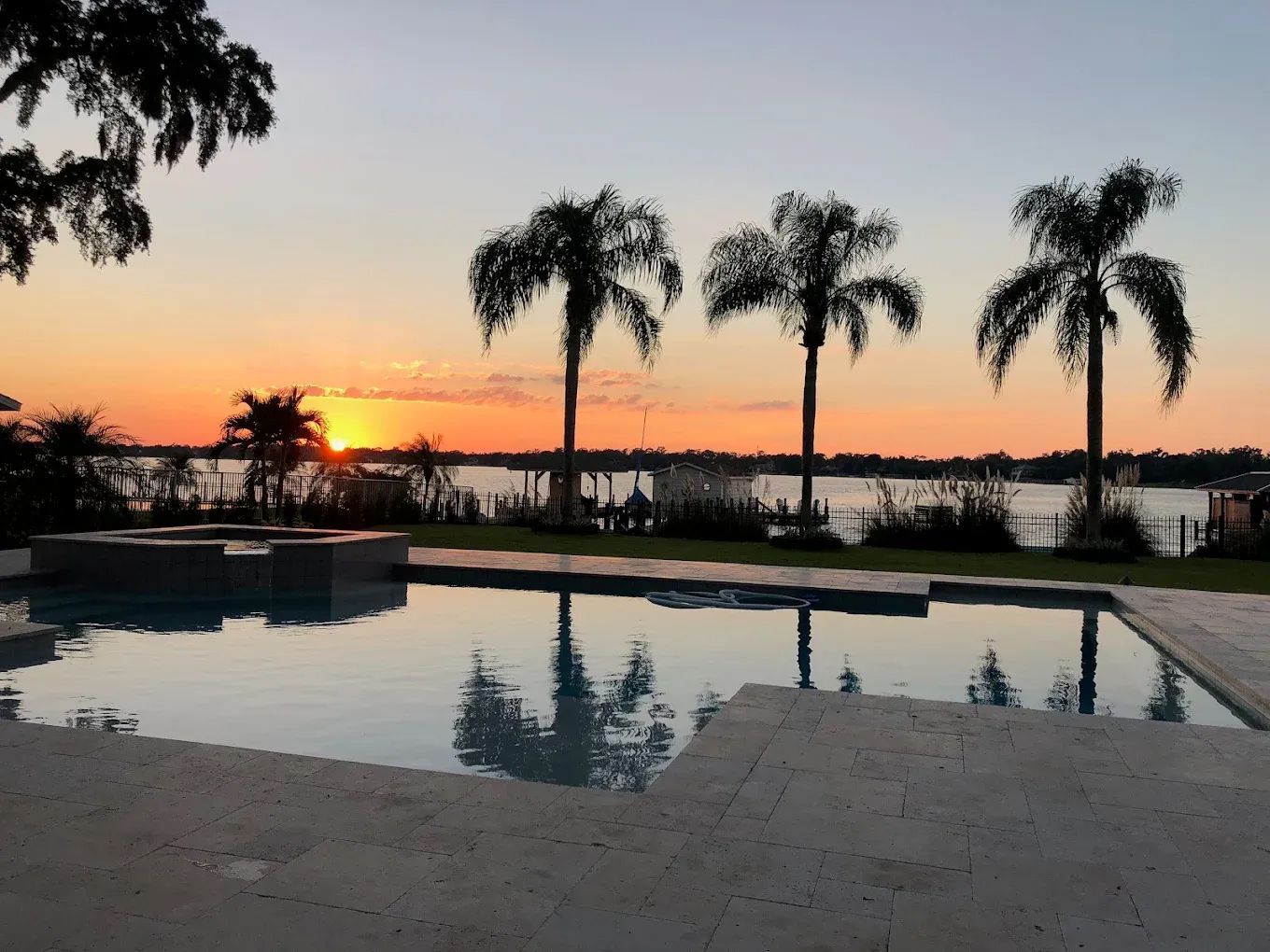 Sunset over water with palm trees and a pool.