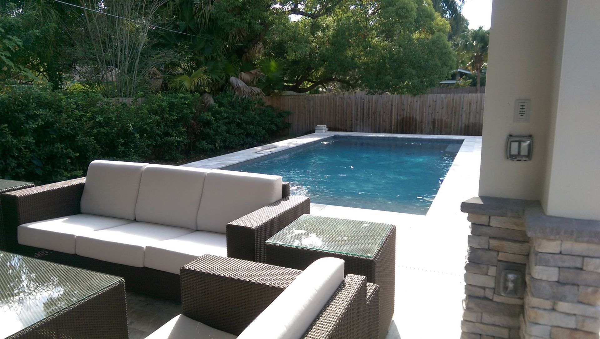 Outdoor patio with pool, modern furniture: brown wicker sofas, glass tables, white concrete, turquoise water, green foliage.