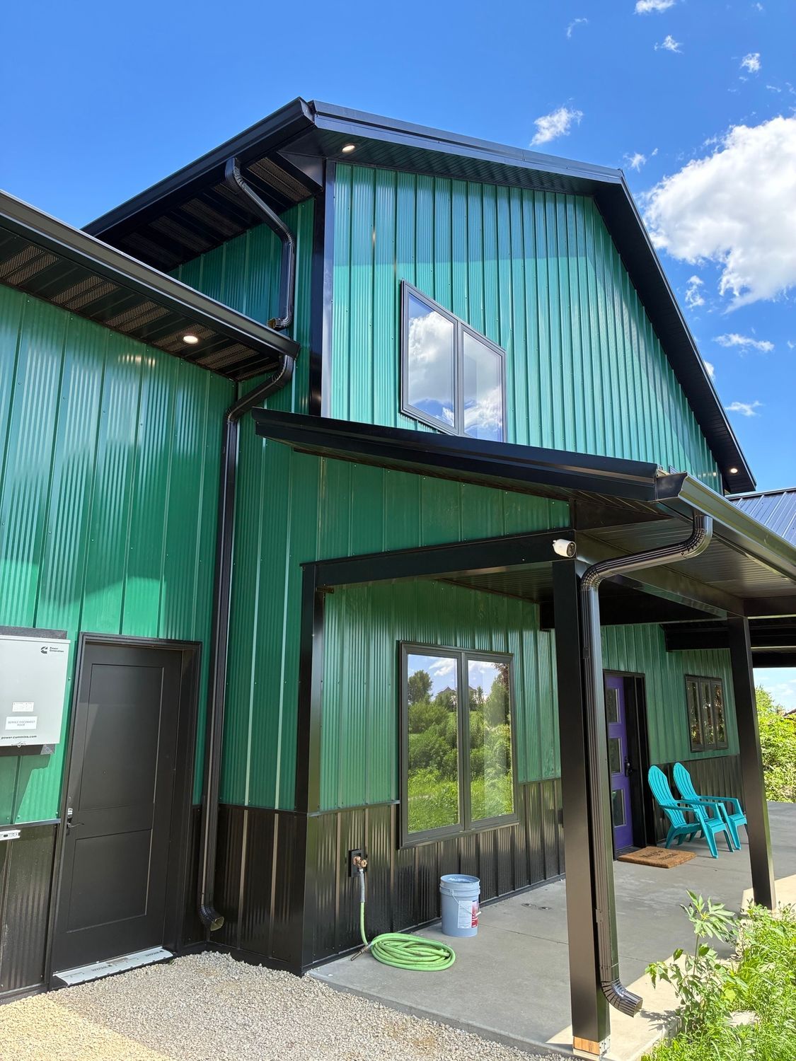 Green metal house exterior with covered porch, black trim, and blue chairs under a bright sky