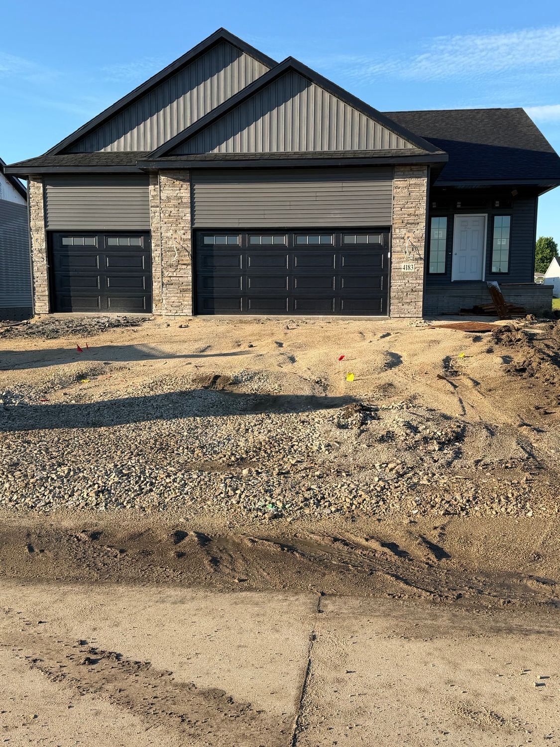 New house with black garage doors, stone columns, and unfinished gravel driveway