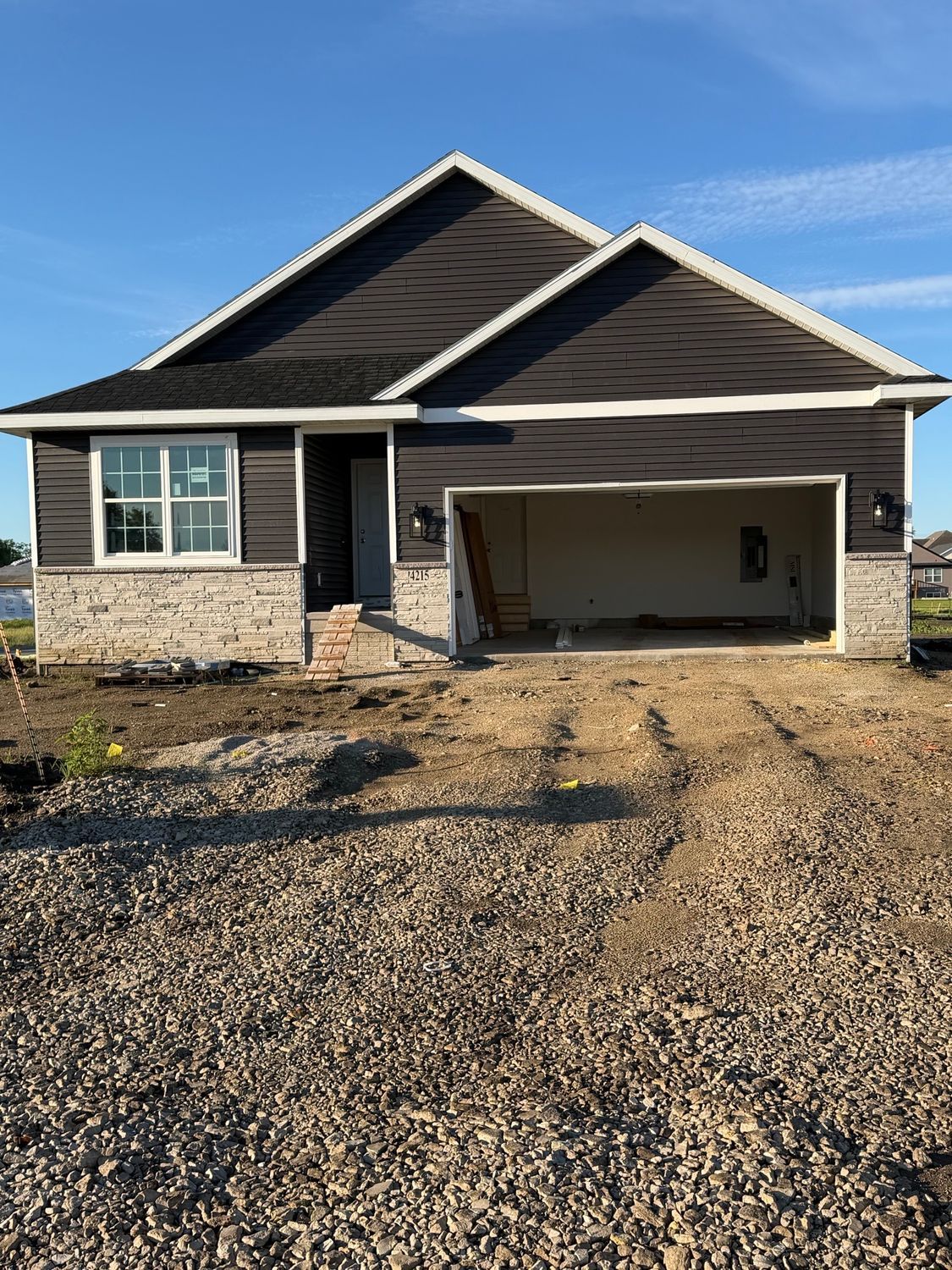 Single-story house with open garage, dark siding, and unfinished gravel driveway under a clear blue sky