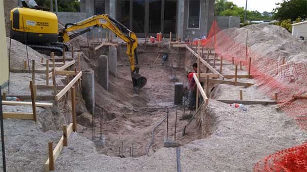 A yellow excavator is digging a hole in the ground in front of a house under construction.