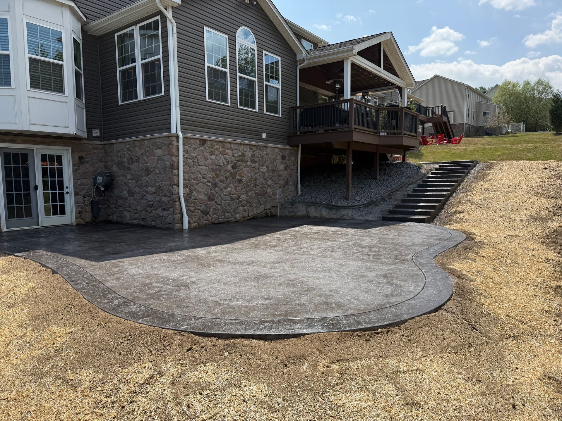 Backyard with a gray concrete patio, steps up a slope, and a deck attached to a brick house.