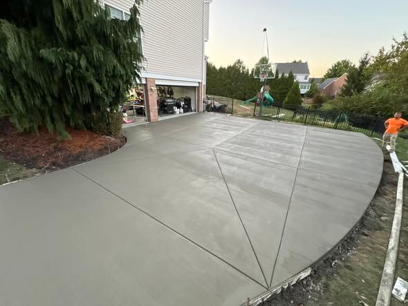 Newly poured concrete driveway and patio. Person works on the edge. House and yard in the background.