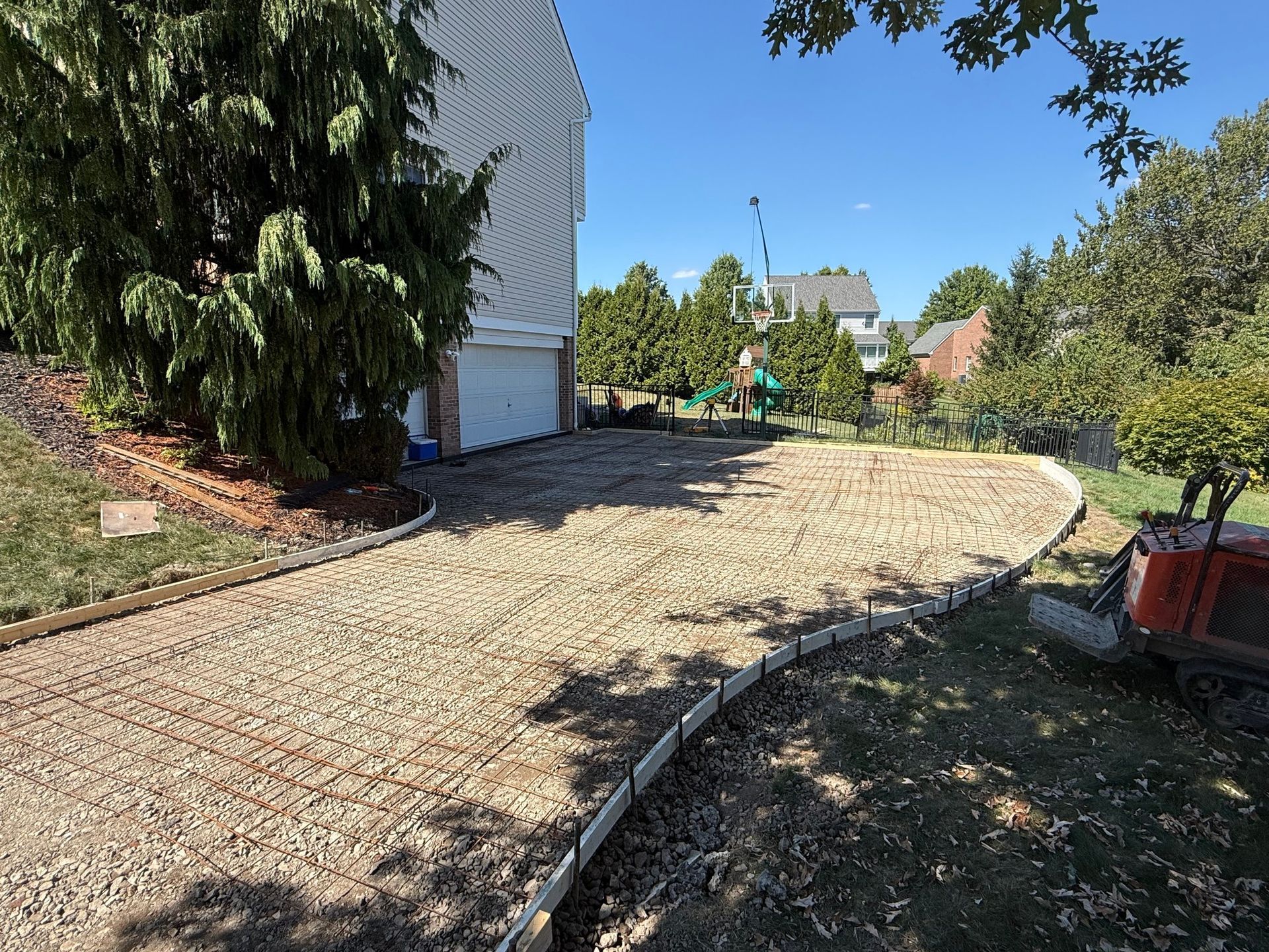 Gravel driveway under construction, next to a house with a garage, in a yard with trees and blue sky.