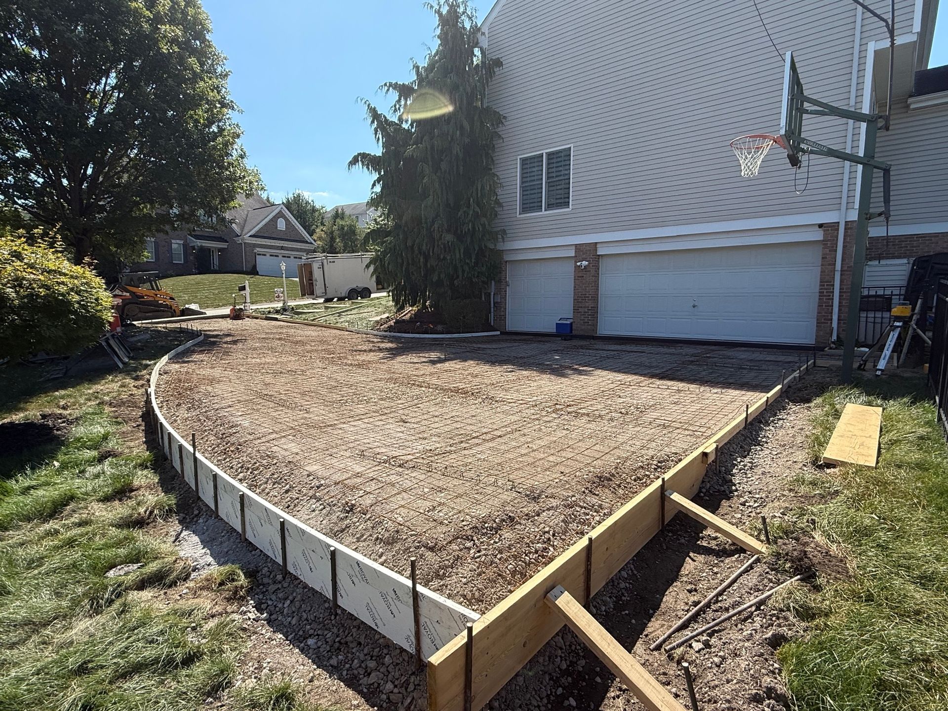 A gravel-filled area framed by wooden forms, prepared for concrete, next to a house with a basketball hoop.