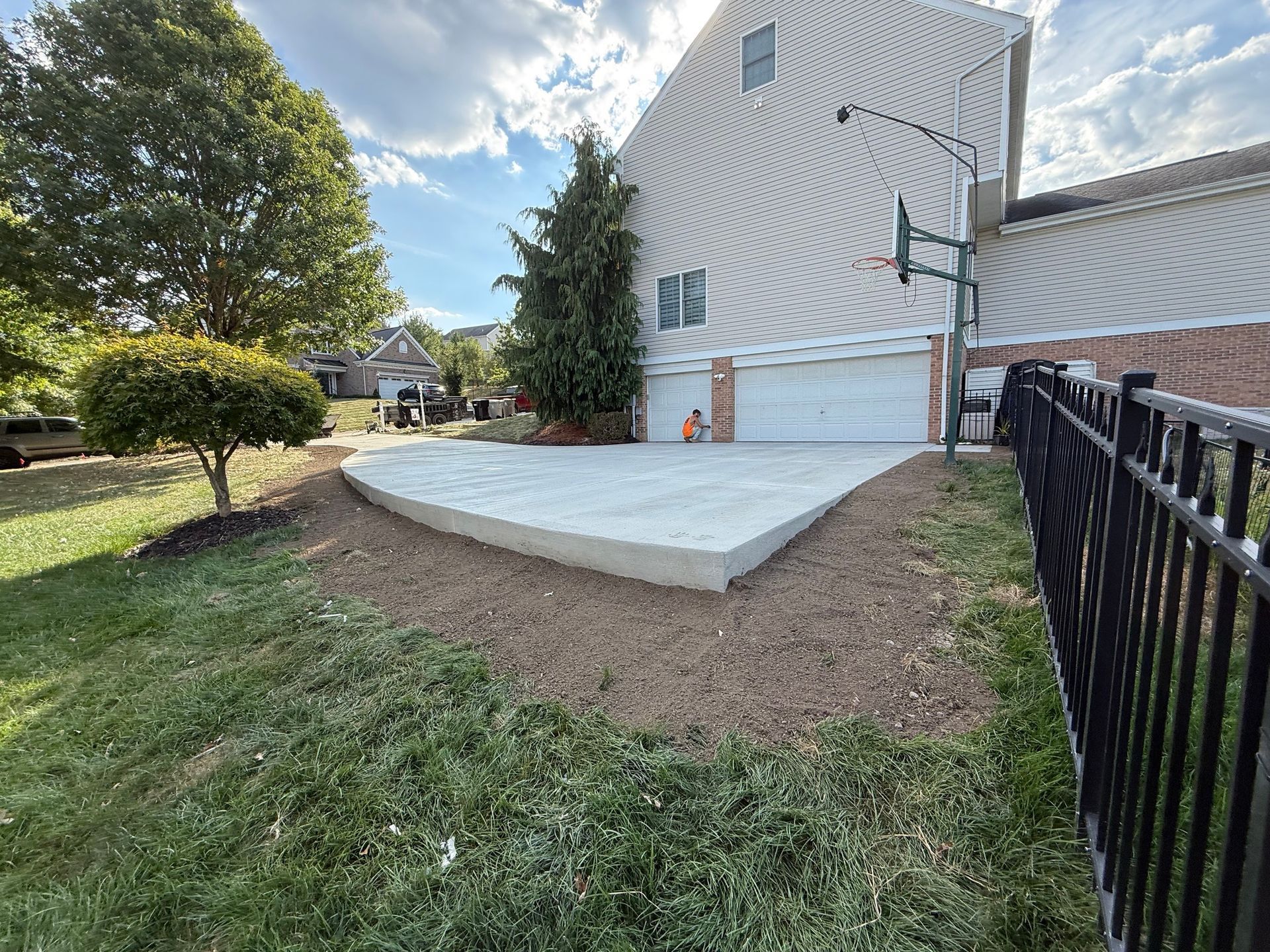 Concrete patio next to a house with a basketball hoop, bordered by lawn and a fence.