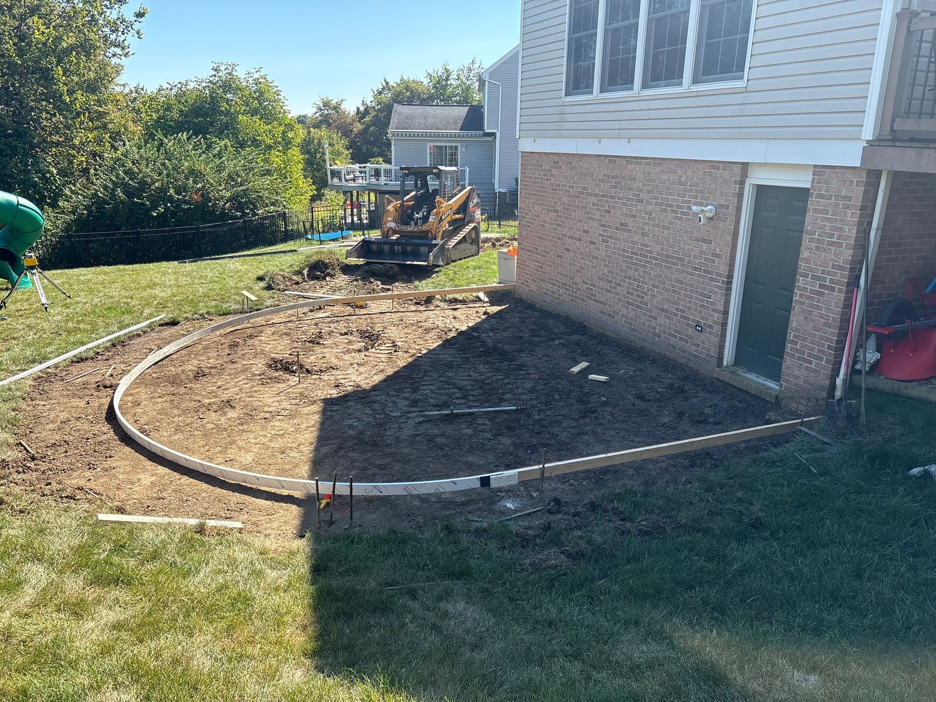 Construction site, a curved concrete border outlines a patio area near a house. A skid steer loader is in the background.