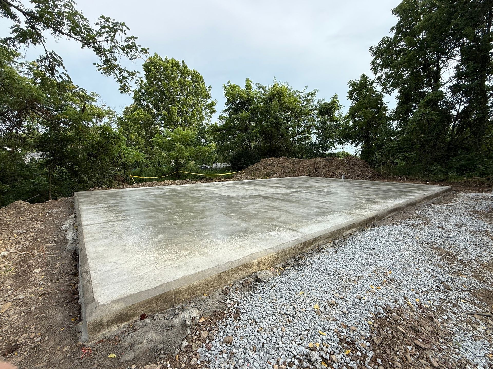 Concrete foundation in outdoor setting, surrounded by gravel and trees. Cloudy sky in background.