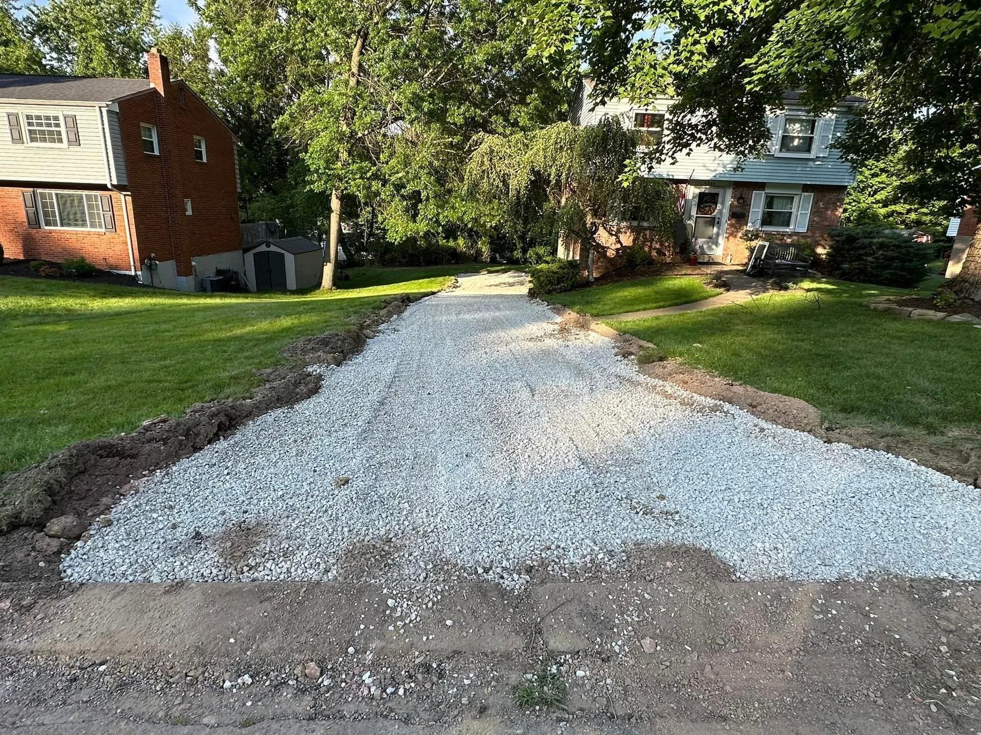 Gravel driveway under construction between two houses, surrounded by grass and trees.