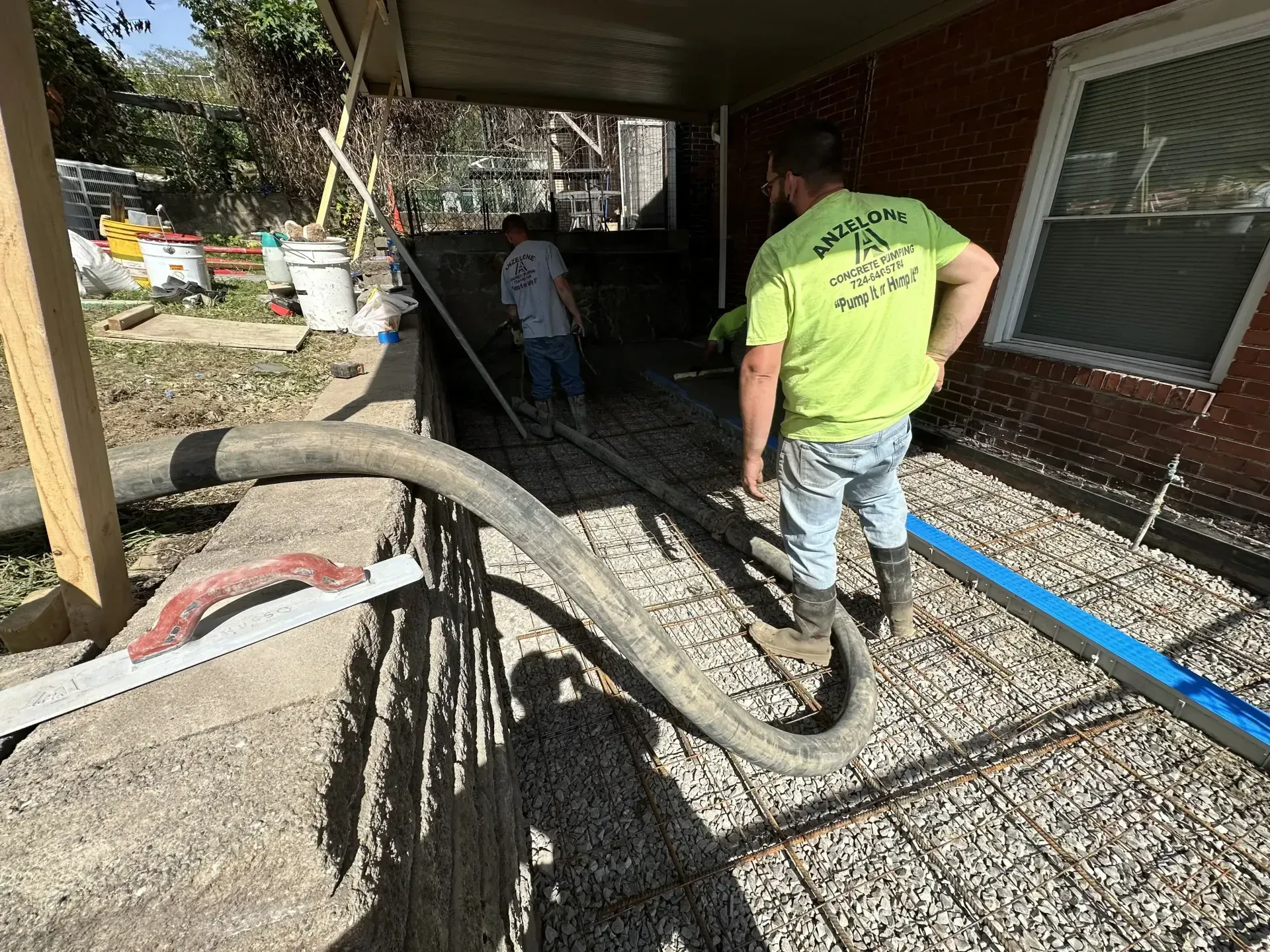 Workers pouring concrete, using hose, on a rebar-laid surface under a porch roof.