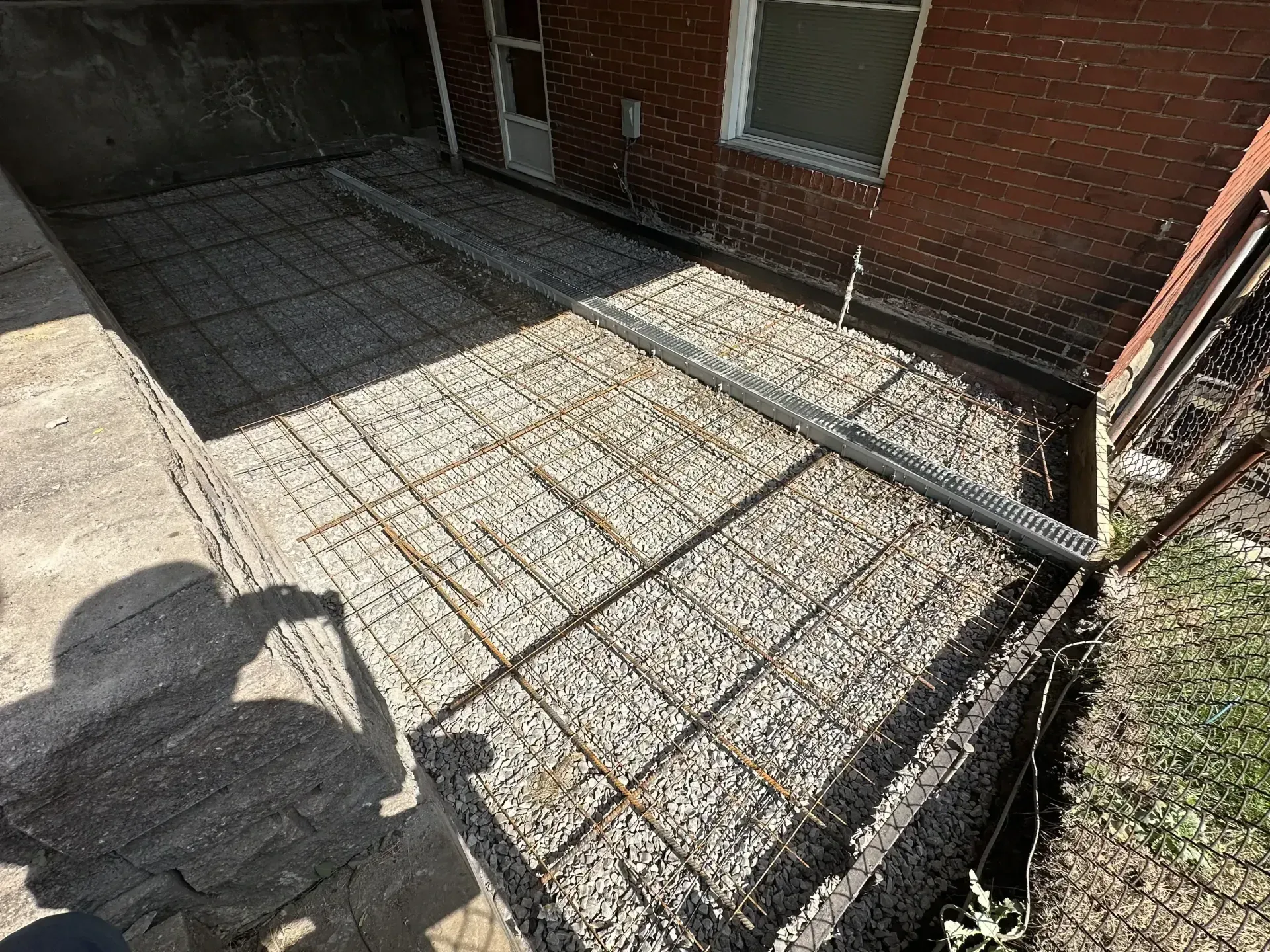 Preparation for concrete slab: rebar grid laid on gravel base next to brick building and wall.