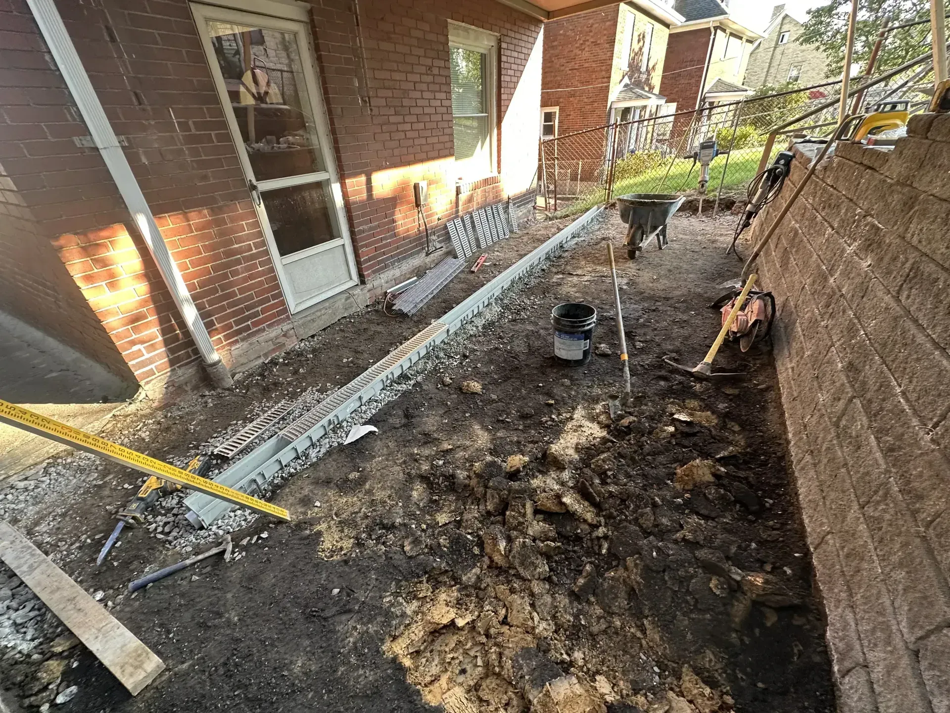 Exterior construction site with dirt, tools, and exposed foundation. A red brick building and concrete wall are present.