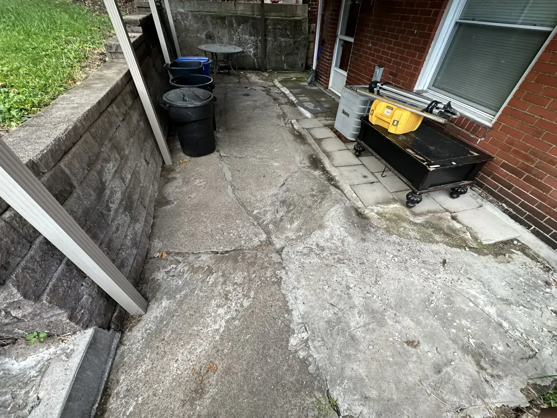Cracked concrete pathway between brick wall, building with window, and retaining wall; trash can, table, and bench.