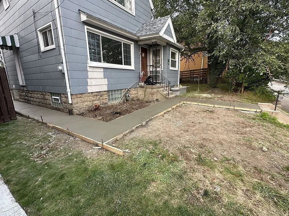 New concrete walkway leading to the front door of a gray house. Green grass and bare earth surround.