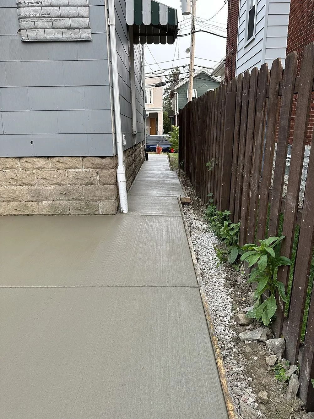 New concrete walkway next to a weathered brown fence and a gray building with a green awning.