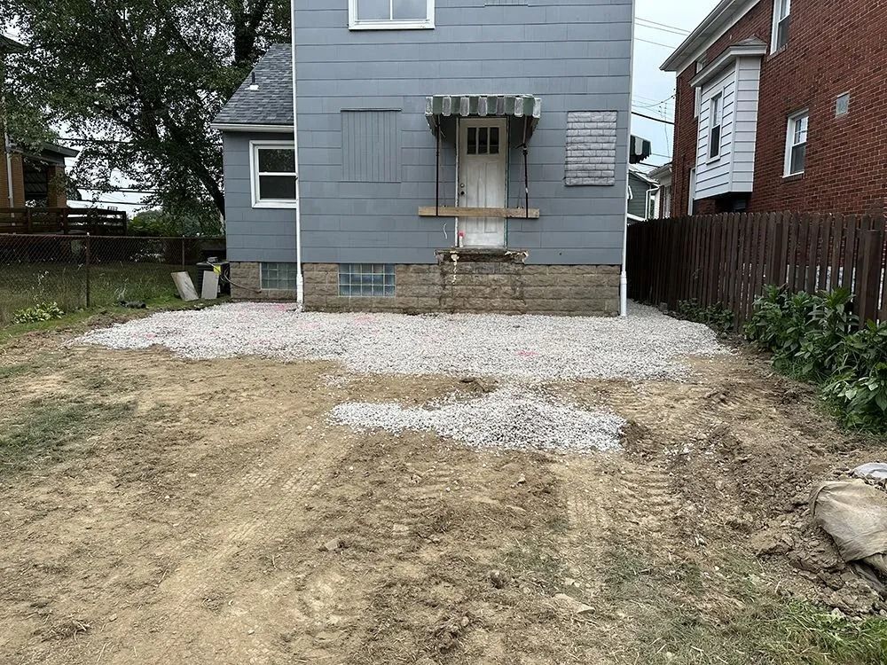 Gravel laid in front of a blue house with a closed door, brown fence, and muddy ground.
