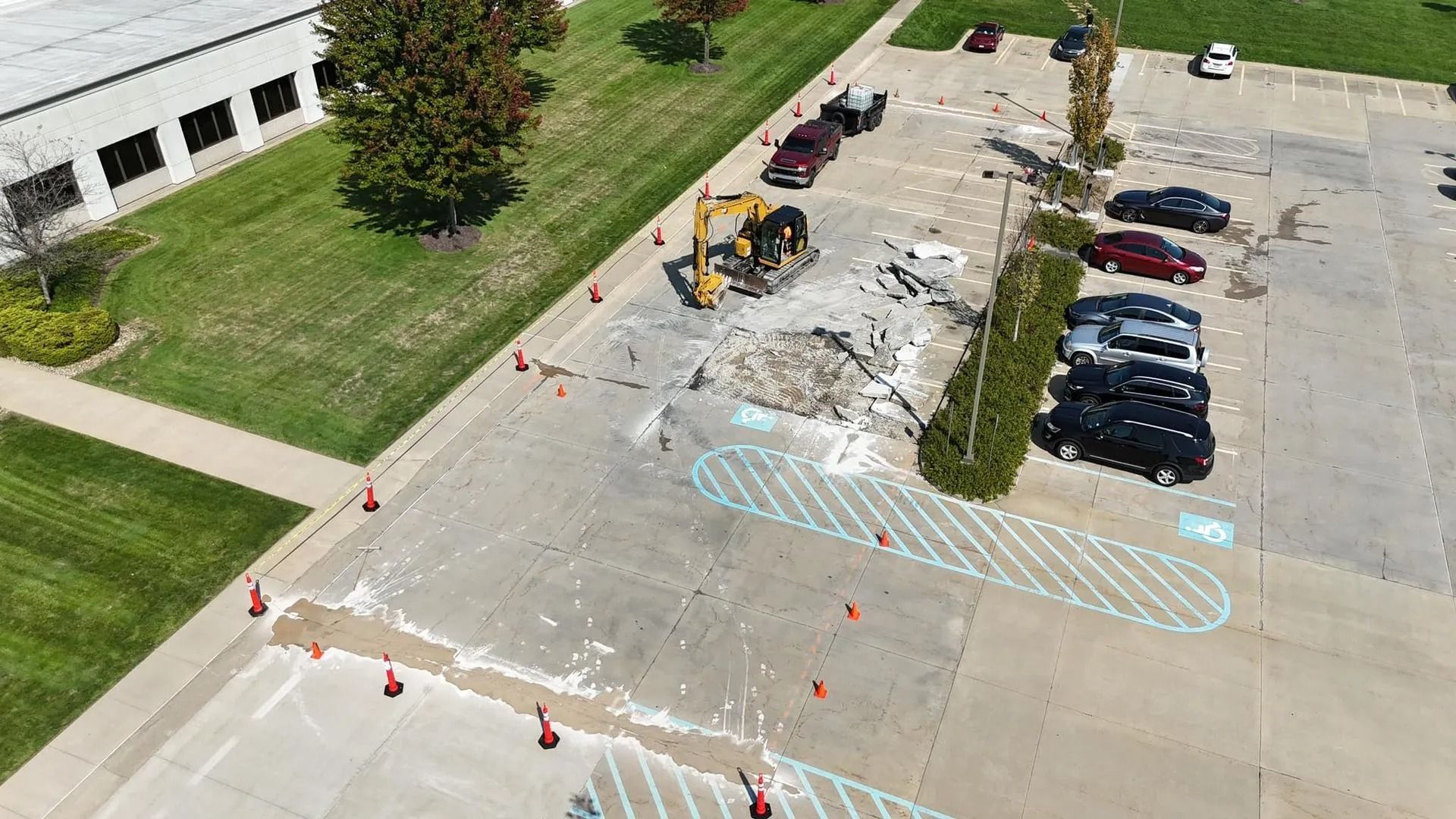 Construction in a parking lot: excavator, debris, orange cones, parked cars, grass, and a building.