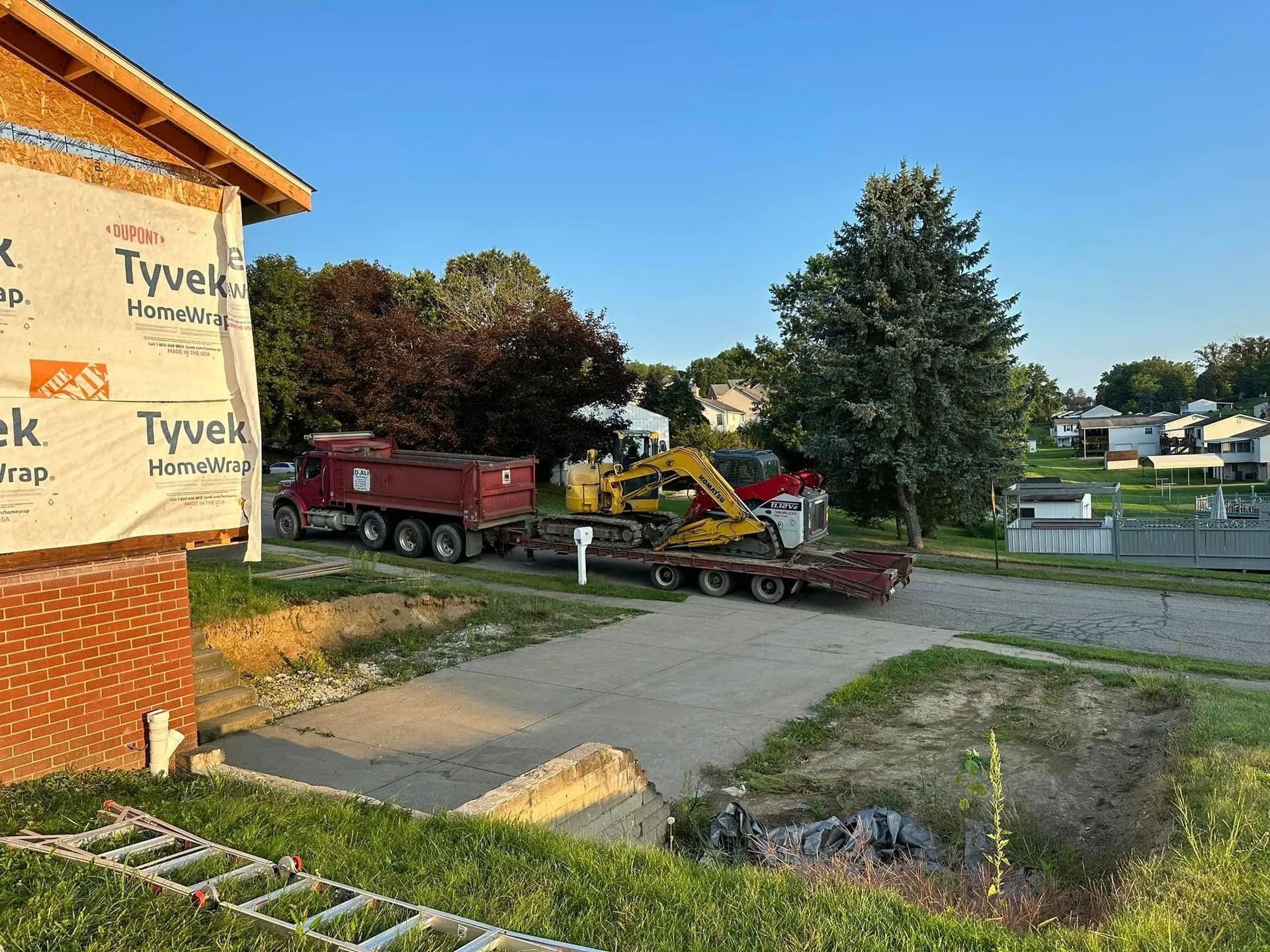 Construction site: a truck with an excavator on a trailer on a concrete driveway near a house under construction.