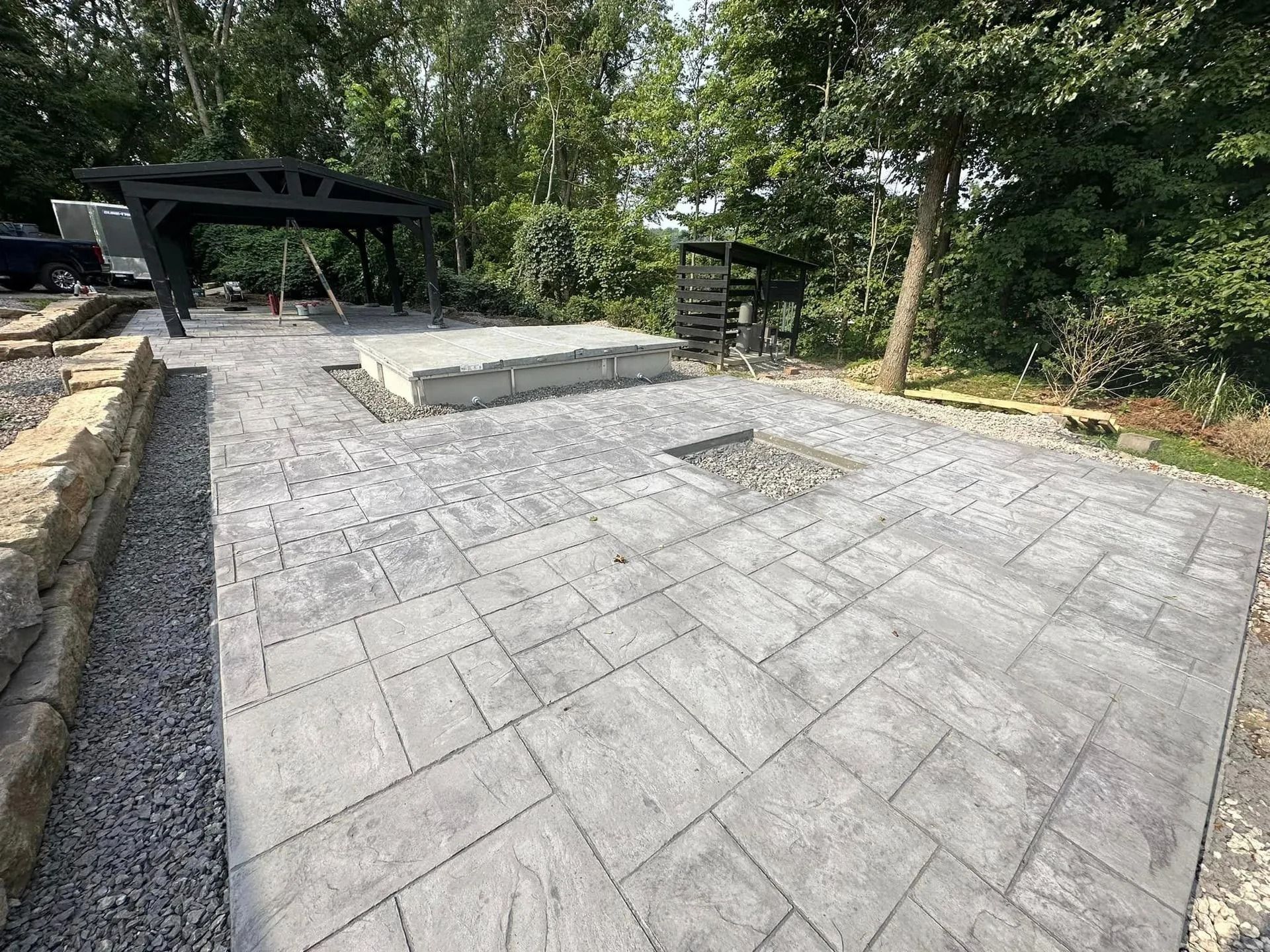 Gray stone patio with pergola, fire pit, and gravel border in a wooded area.