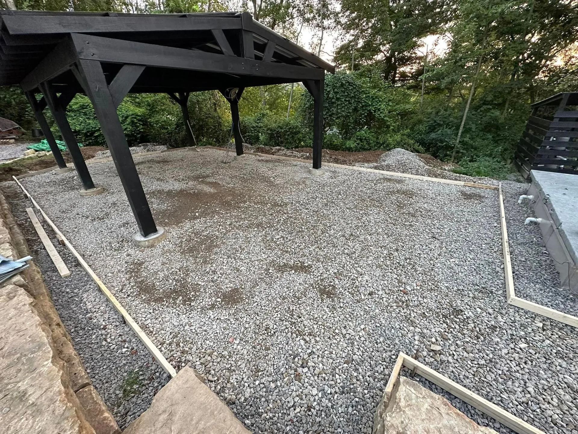 Gravel-covered patio area under a black wooden pergola frame, bordered by wooden edging.
